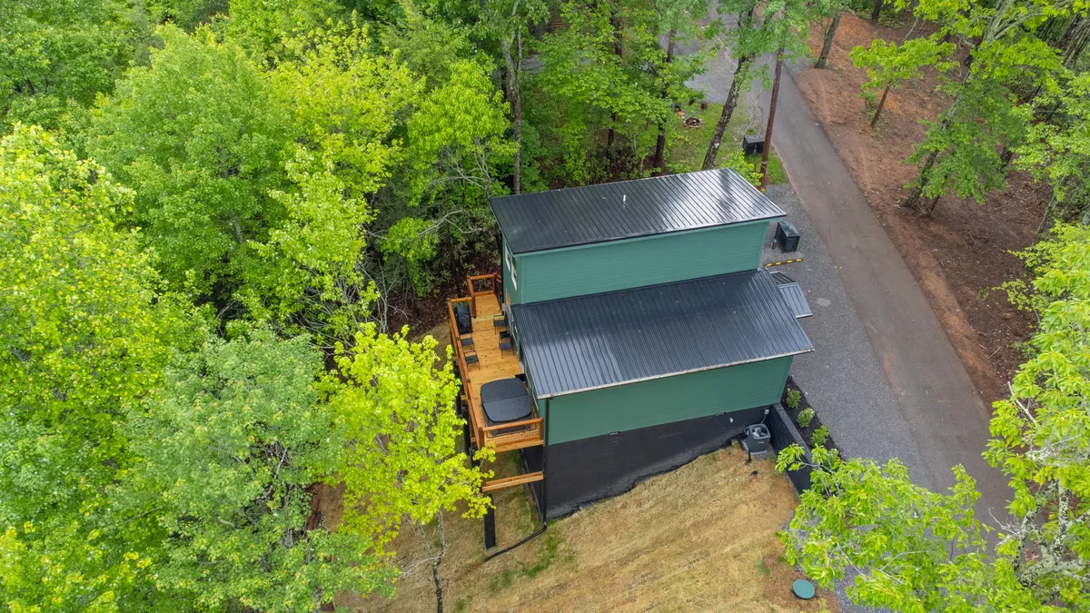 A modern green cabin sits amidst vibrant trees, with a spacious wooden deck inviting outdoor relaxation. The deck offers views of the natural landscape, capturing the peaceful essence of the Smokies.
