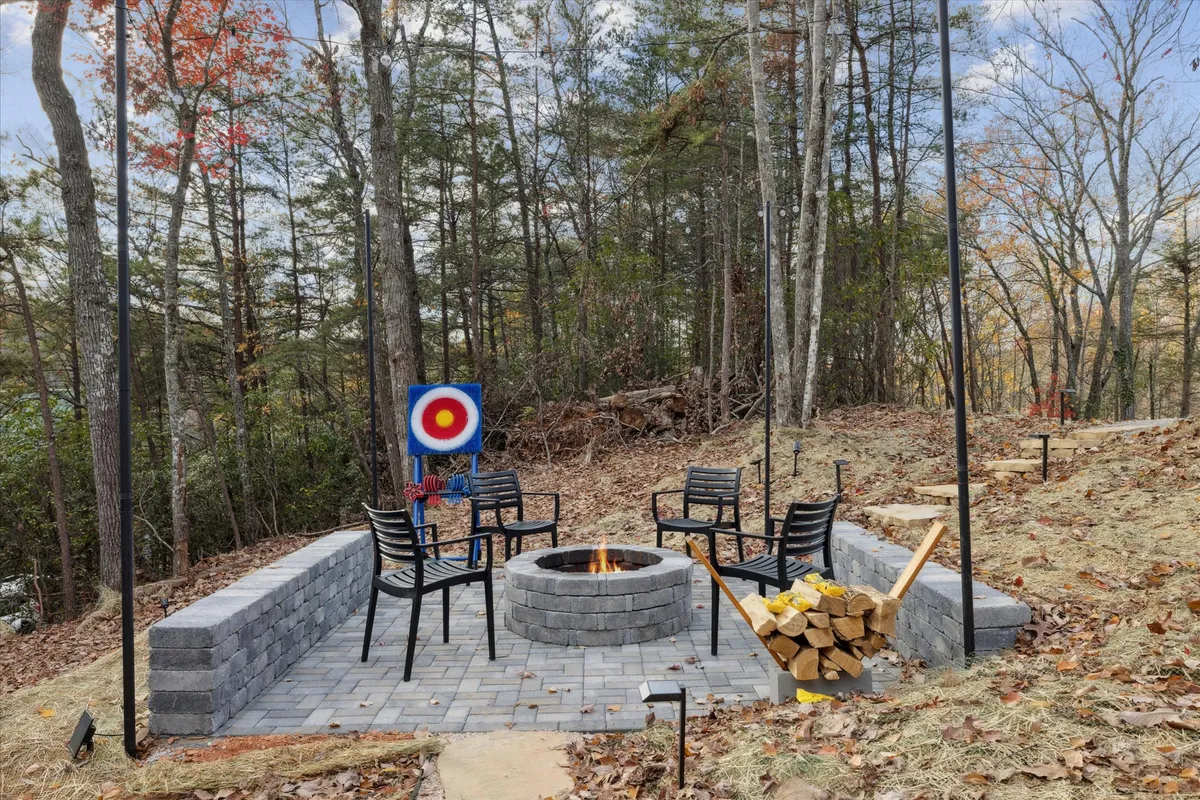 An inviting exterior scene featuring a fire pit, surrounding seating, an archery (or axe-throwing) target, and a wood stack. The environment is designed for outdoor fun, group gatherings, and social interaction around the fire.