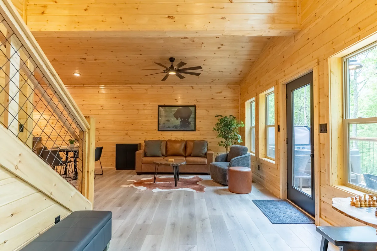 This image features a cozy living area with a sofa, armchairs, a coffee table, and large windows that frame views of the surrounding woods. The artwork on the wall and the soft lighting give the room a welcoming and comfortable feel, perfect for relaxing or socializing.