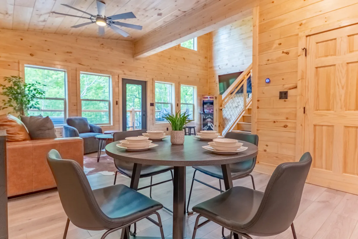 The photo shows an open-concept space with a dining table set for a meal, a comfortable seating area, a wooden staircase, and large windows that let in ample natural light. The space is airy, warm, and inviting, ideal for both meals and casual gatherings.