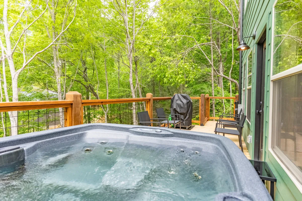 An exterior deck scene featuring a black outdoor grill, hot tub, wooden railing, green trees, and outdoor chairs. The grill is positioned for easy barbecuing just steps from the house, and the space radiates a serene yet social vibe ideal for evening gatherings.