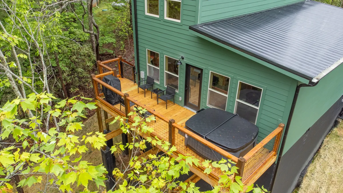 This exterior shot showcases the green exterior of the cabin, a spacious wooden deck, inviting hot tubs, and a seating area. The scene captures a tranquil, welcoming mood perfect for relaxing or entertaining guests after mountain adventures.